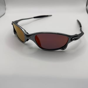 May include: A pair of gray sunglasses with a sleek, modern design. The lenses are a gradient of orange and red, and the frames are a matte gray. The temples are black and curve around the ears. The sunglasses are set against a white background.