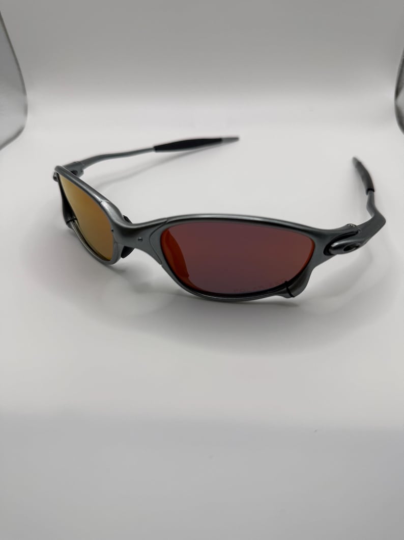 May include: A pair of gray sunglasses with a sleek, modern design. The lenses are a gradient of orange and red, and the frames are a matte gray. The temples are black and curve around the ears. The sunglasses are set against a white background.