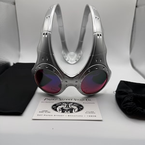 Könnte beinhalten: Silberfarbene Steampunk-Brille mit rot-violetten Gläsern. Die Brille hat ein einzigartiges, industrielles Design mit sichtbaren Nieten und einem Steg. Eine Visitenkarte und zwei schwarze Beutel sind ebenfalls im Bild.