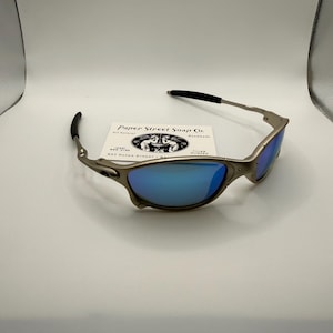 May include: Silver-framed sunglasses with blue lenses. The sunglasses have black earpieces and a small business card is behind them. The card reads "Paper Street Soap Co." with a logo of two figures.