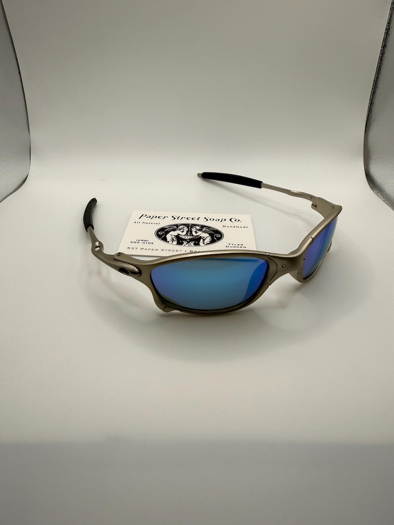 May include: Silver-framed sunglasses with blue lenses. The sunglasses have black earpieces and a small business card is behind them. The card reads "Paper Street Soap Co." with a logo of two figures.