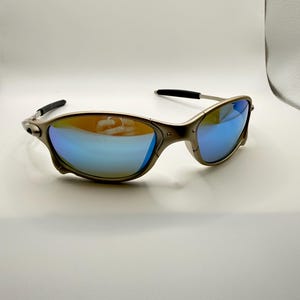 May include: A pair of sunglasses with a metallic frame and blue mirrored lenses. The lenses reflect a gradient of colors, and the temples are black. The sunglasses are designed for outdoor wear and offer UV protection.