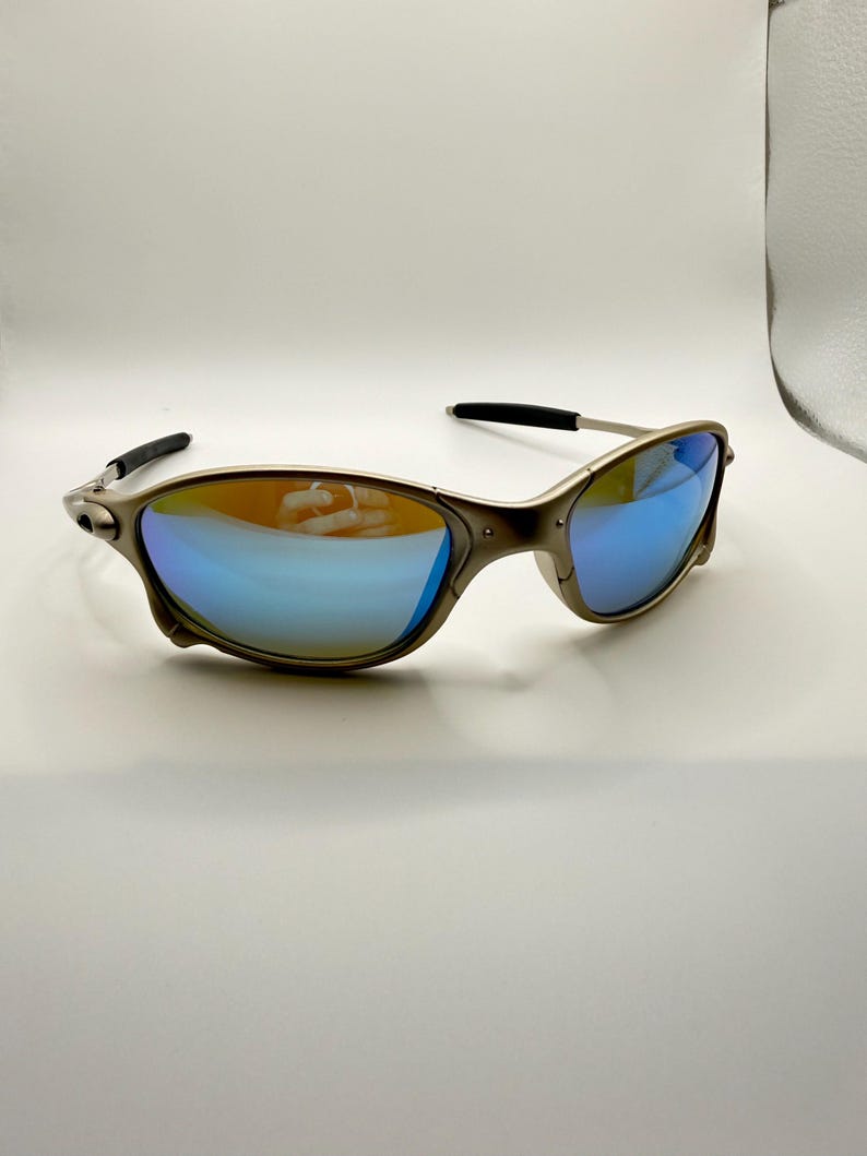 May include: A pair of sunglasses with a metallic frame and blue mirrored lenses. The lenses reflect a gradient of colors, and the temples are black. The sunglasses are designed for outdoor wear and offer UV protection.