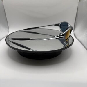 May include: Silver-framed sunglasses with blue and yellow lenses rest on a reflective black surface. The sunglasses have a sleek, modern design with a curved frame and dark temples. The lenses reflect the surrounding environment.