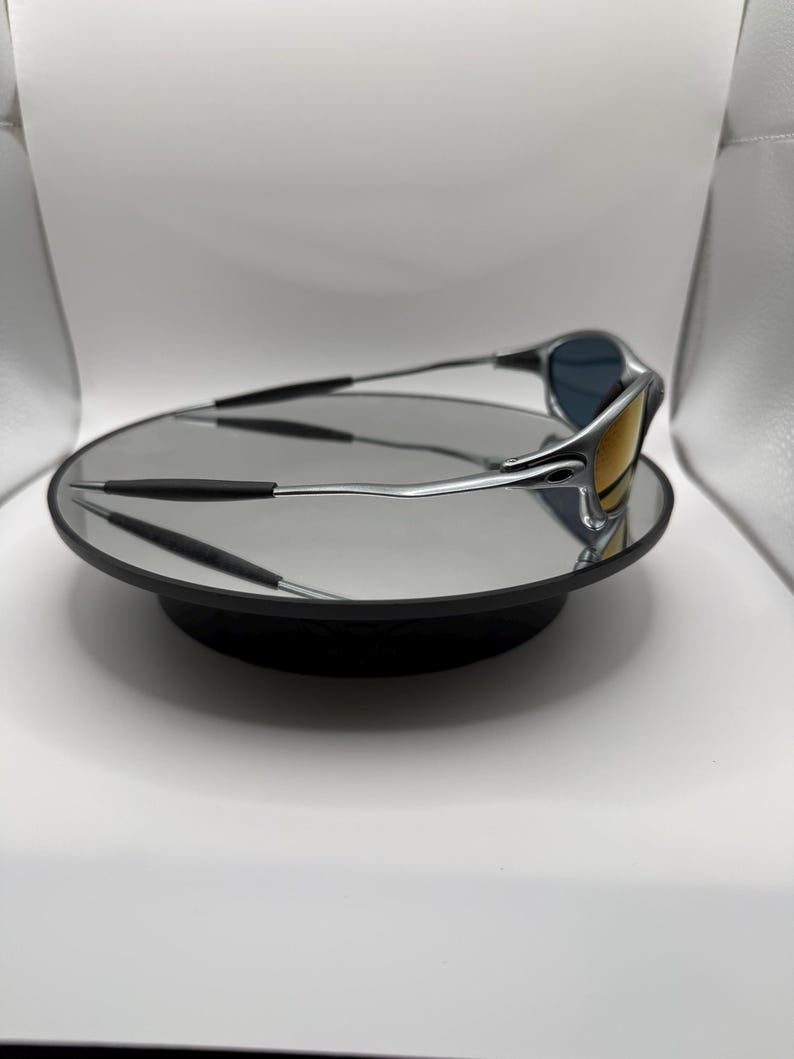 May include: Silver-framed sunglasses with blue and yellow lenses rest on a reflective black surface. The sunglasses have a sleek, modern design with a curved frame and dark temples. The lenses reflect the surrounding environment.