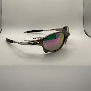 May include: A pair of sunglasses with a metallic gray frame and mirrored lenses that reflect pink, green, and blue hues. The temples have black rubber grips. The sunglasses are on a white surface.