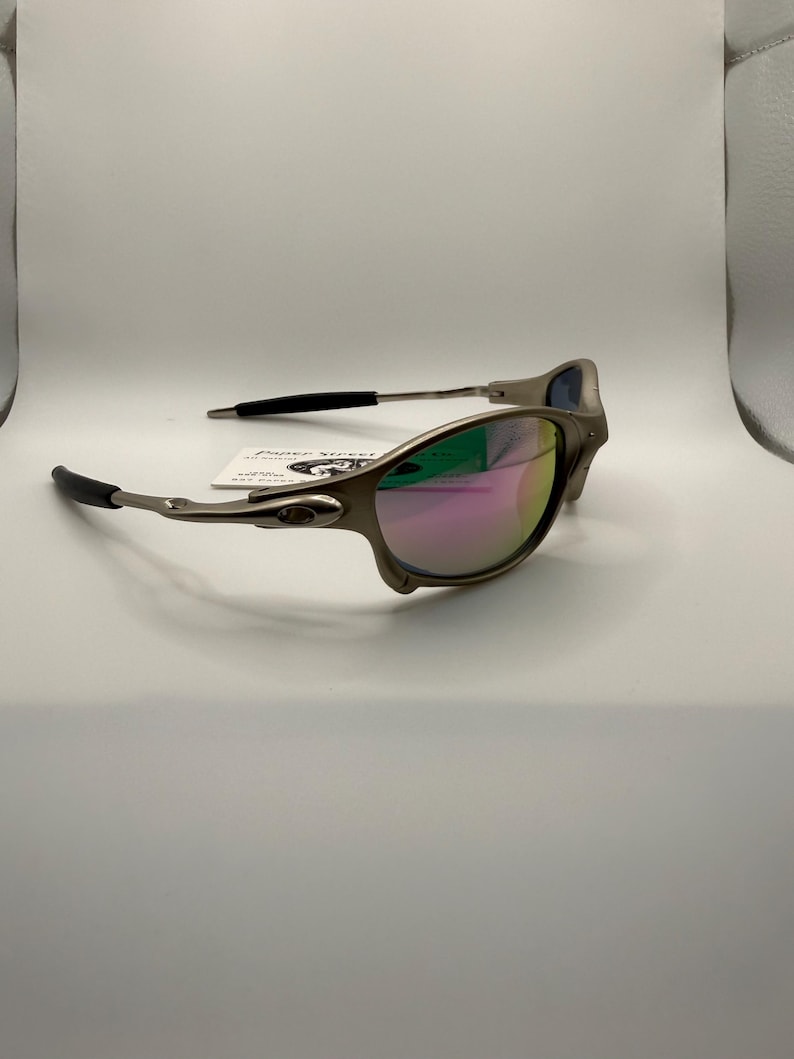 May include: A pair of sunglasses with a metallic gray frame and mirrored lenses that reflect pink, green, and blue hues. The temples have black rubber grips. The sunglasses are on a white surface.