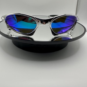 Op de afbeelding: Zilverkleurige zonnebril met blauwe spiegelglazen. De zonnebril wordt gepresenteerd op een klein, rond, spiegelend oppervlak. De glazen reflecteren een spectrum aan kleuren, wat een levendige uitstraling cre&euml;ert. Het montuur heeft een strak, modern design.