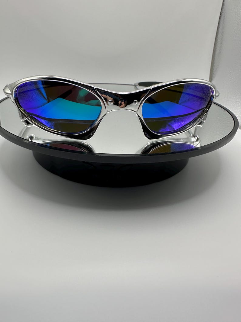 Op de afbeelding: Zilverkleurige zonnebril met blauwe spiegelglazen. De zonnebril wordt gepresenteerd op een klein, rond, spiegelend oppervlak. De glazen reflecteren een spectrum aan kleuren, wat een levendige uitstraling cre&euml;ert. Het montuur heeft een strak, modern design.