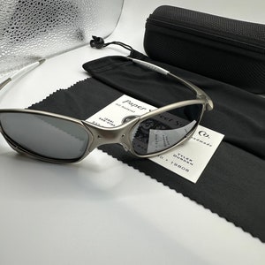 May include: Silver-framed sunglasses with dark lenses, a black carrying case, and a black microfiber cleaning cloth. The sunglasses have a sleek, modern design. A white tag with the text "Paper Street Co." is also present.