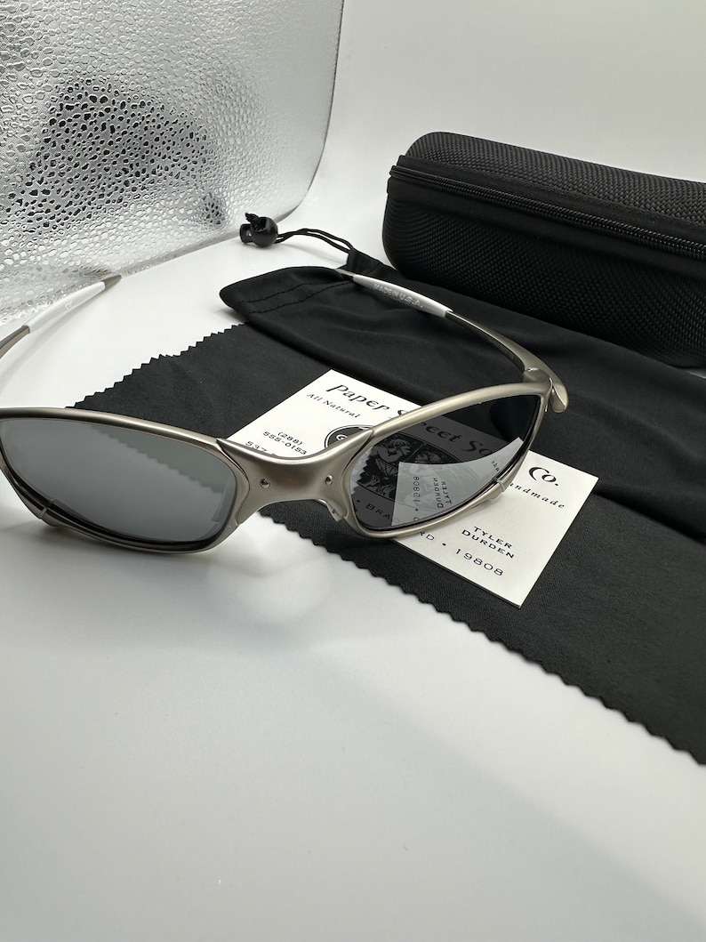 May include: Silver-framed sunglasses with dark lenses, a black carrying case, and a black microfiber cleaning cloth. The sunglasses have a sleek, modern design. A white tag with the text "Paper Street Co." is also present.