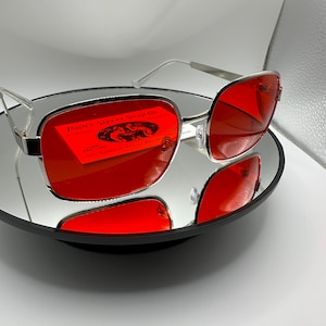 May include: Silver framed sunglasses with red lenses. The lenses reflect a business card for Paper Street Soap Co. with the text "All Natural" and "1281 Collins 587 Paper Street, Bradenton, FL 34205".