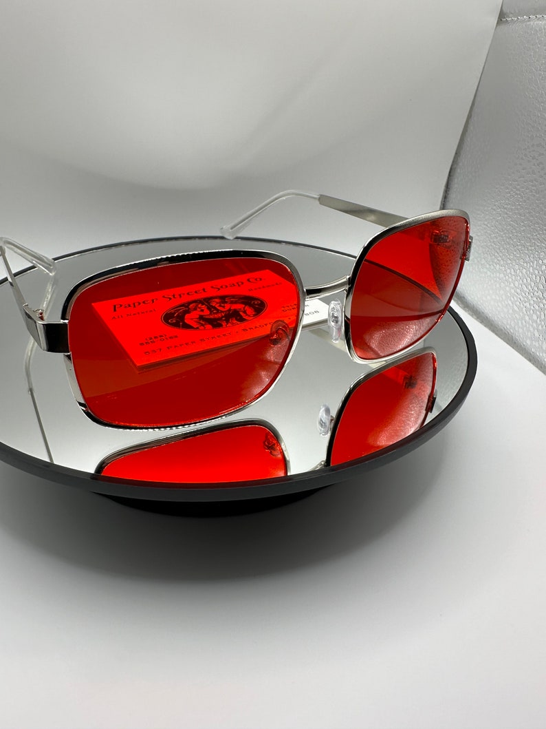 May include: Silver framed sunglasses with red lenses. The lenses reflect a business card for Paper Street Soap Co. with the text "All Natural" and "1281 Collins 587 Paper Street, Bradenton, FL 34205".