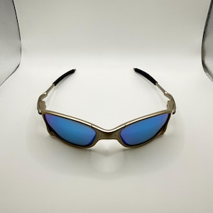 May include: Sunglasses with a metallic frame and blue lenses. The sunglasses have a sleek, modern design with a curved frame and black earpieces. The lenses reflect a bright blue color, offering a stylish look.