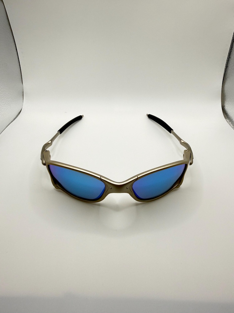 May include: Sunglasses with a metallic frame and blue lenses. The sunglasses have a sleek, modern design with a curved frame and black earpieces. The lenses reflect a bright blue color, offering a stylish look.