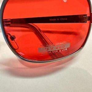 Puede incluir: Gafas de sol con montura plateada y lentes rojas. Las gafas tienen una etiqueta que dice "100% Eye Protection UV 400 Against UVA & UVB" y "Made in China".