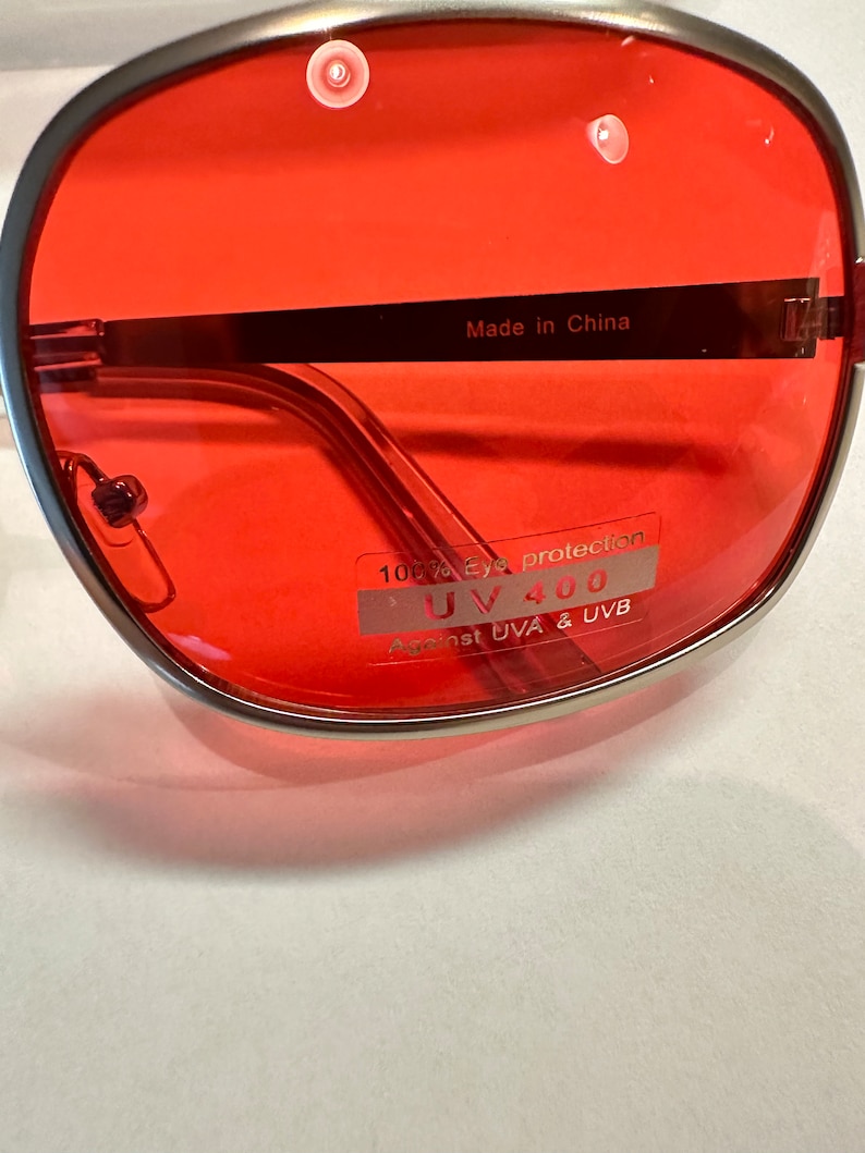 Puede incluir: Gafas de sol con montura plateada y lentes rojas. Las gafas tienen una etiqueta que dice "100% Eye Protection UV 400 Against UVA & UVB" y "Made in China".
