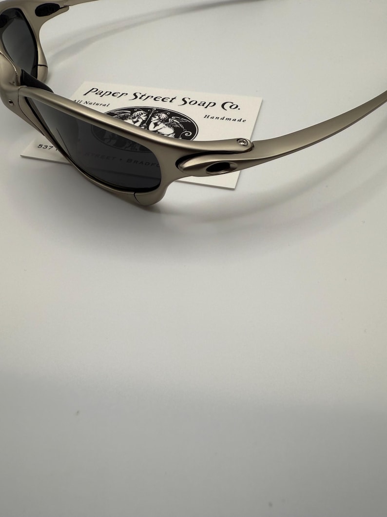 Peut inclure: Lunettes de soleil grises m&eacute;tallis&eacute;es avec des verres fonc&eacute;s. Les lunettes de soleil reposent sur une carte blanche avec l'inscription "Paper Street Soap Co." et une illustration de deux anges. La monture a un design &eacute;l&eacute;gant et moderne.