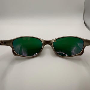 May include: A pair of sunglasses with a metallic gray frame and green lenses. The lenses reflect light, and the frame has a sleek, modern design. The sunglasses are designed for eye protection and fashion.