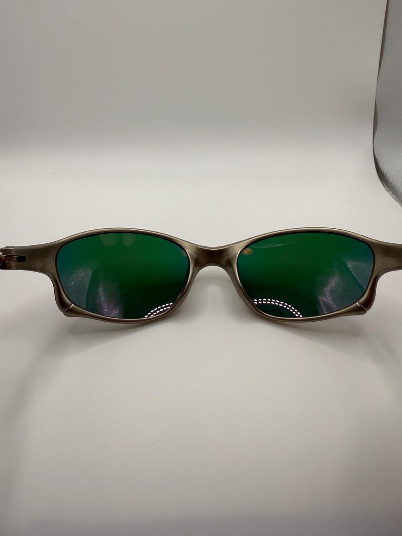 May include: A pair of sunglasses with a metallic gray frame and green lenses. The lenses reflect light, and the frame has a sleek, modern design. The sunglasses are designed for eye protection and fashion.