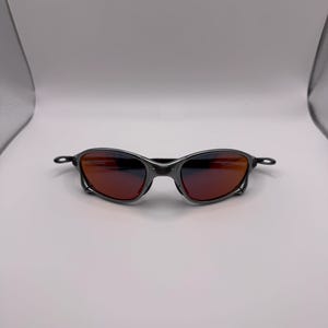 May include: A pair of sunglasses with a dark gray metal frame and reddish-orange lenses. The sunglasses have a sleek, modern design, suitable for outdoor activities. The lenses provide protection from the sun's glare.