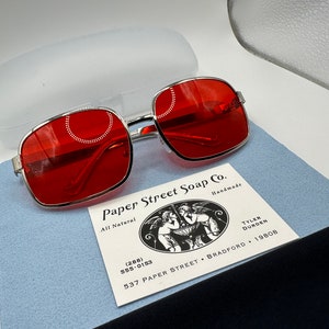 May include: Red square-shaped sunglasses with a silver metal frame. The sunglasses are sitting on a blue surface next to a business card for Paper Street Soap Co. The business card has a black and white illustration of two angels holding a bowl. The business card includes the phone number (288) 555-0153, the address 537 Paper Street, Bradford, and the year 19808.