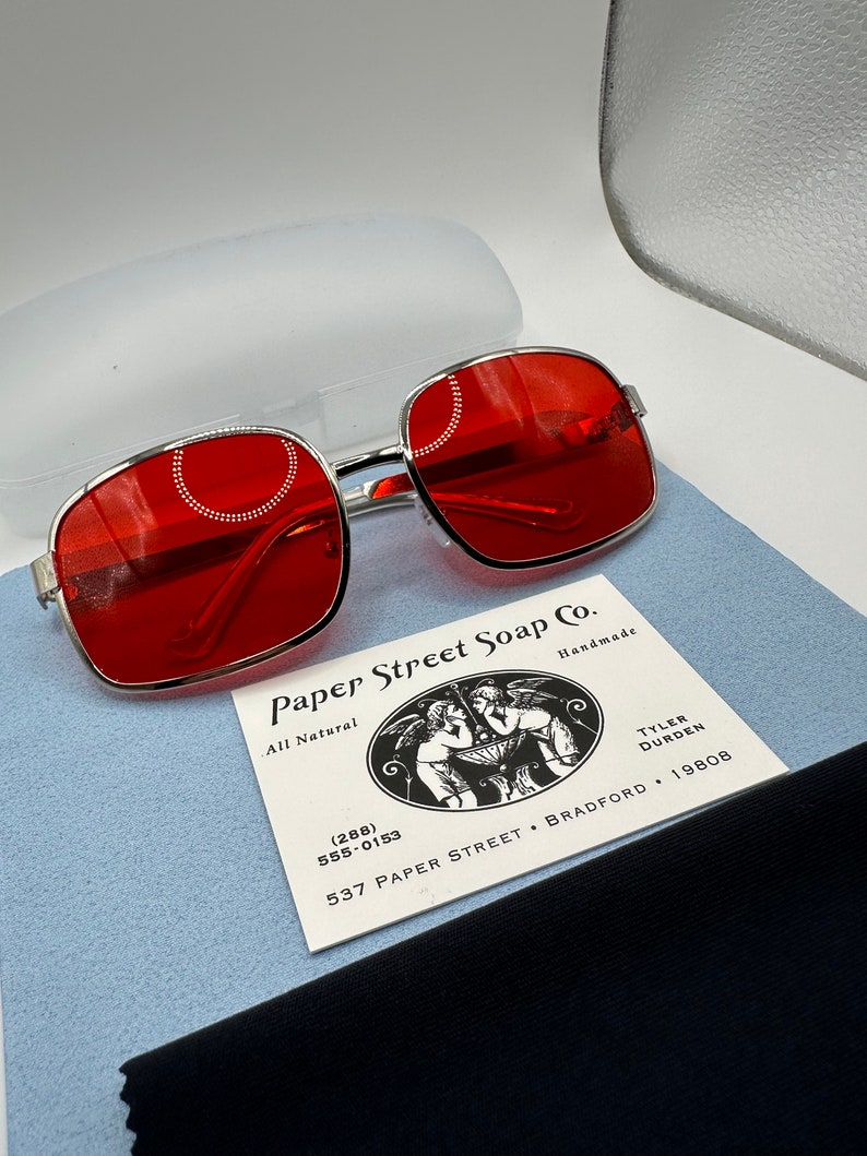 May include: Red square-shaped sunglasses with a silver metal frame. The sunglasses are sitting on a blue surface next to a business card for Paper Street Soap Co. The business card has a black and white illustration of two angels holding a bowl. The business card includes the phone number (288) 555-0153, the address 537 Paper Street, Bradford, and the year 19808.
