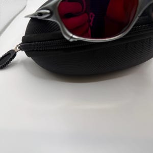 May include: Silver-framed sunglasses with red lenses rest on a black zippered case. The sunglasses have a sleek, modern design. The case is made of a textured material.