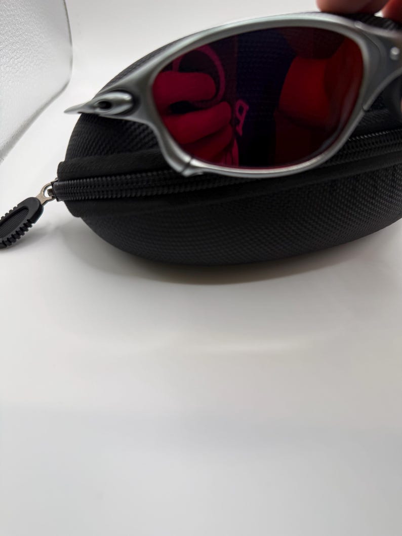May include: Silver-framed sunglasses with red lenses rest on a black zippered case. The sunglasses have a sleek, modern design. The case is made of a textured material.