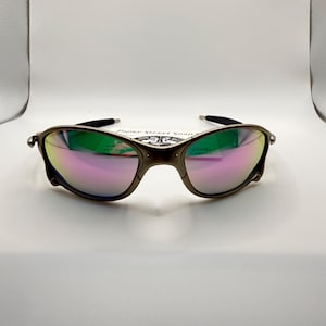 May include: A pair of sunglasses with a dark metallic frame and iridescent lenses reflecting green and pink hues. The sunglasses are set against a white background.
