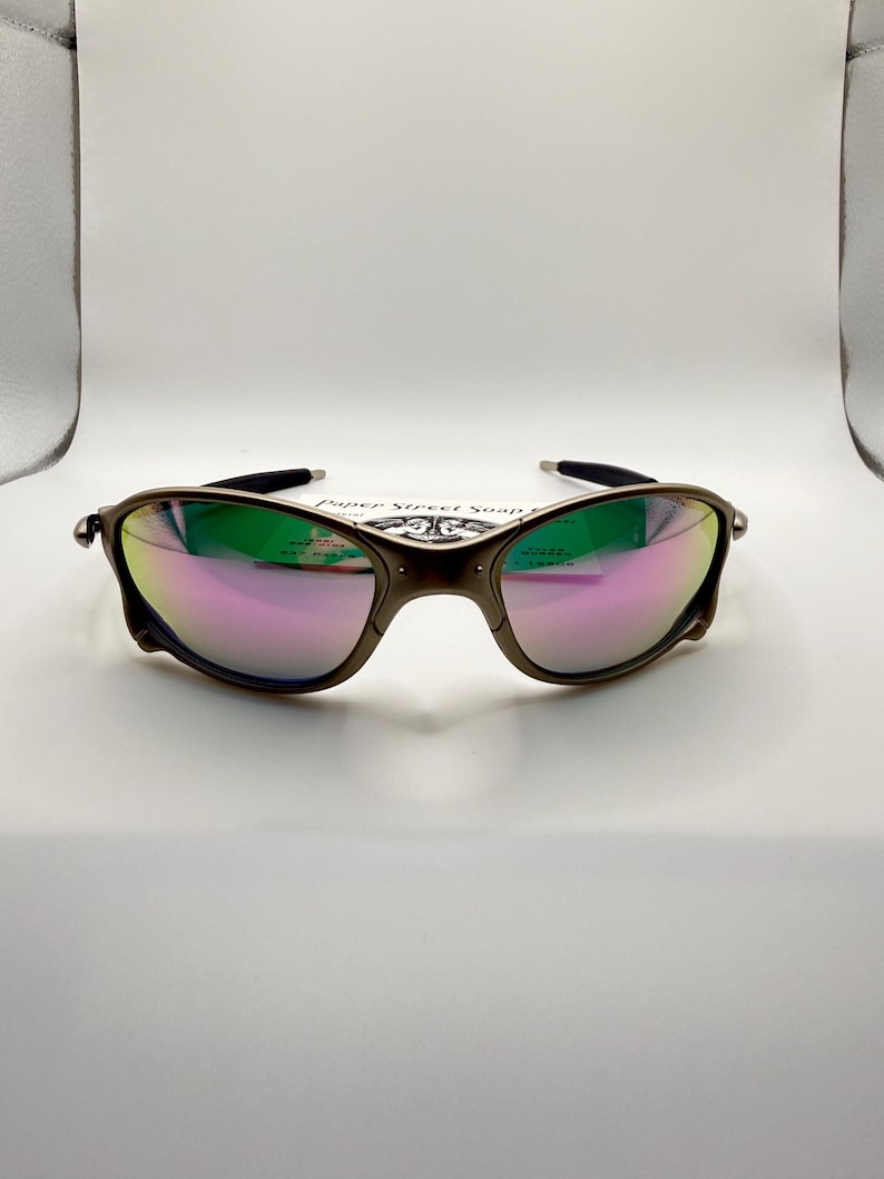 May include: A pair of sunglasses with a dark metallic frame and iridescent lenses reflecting green and pink hues. The sunglasses are set against a white background.
