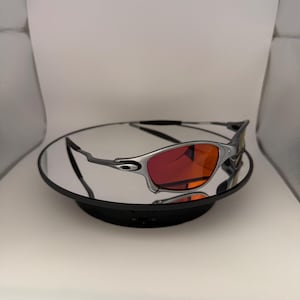 May include: Silver-framed sunglasses with vibrant red lenses. The sunglasses are displayed on a reflective surface, showcasing their sleek design. The lenses offer a striking contrast against the silver frame, creating a modern aesthetic.