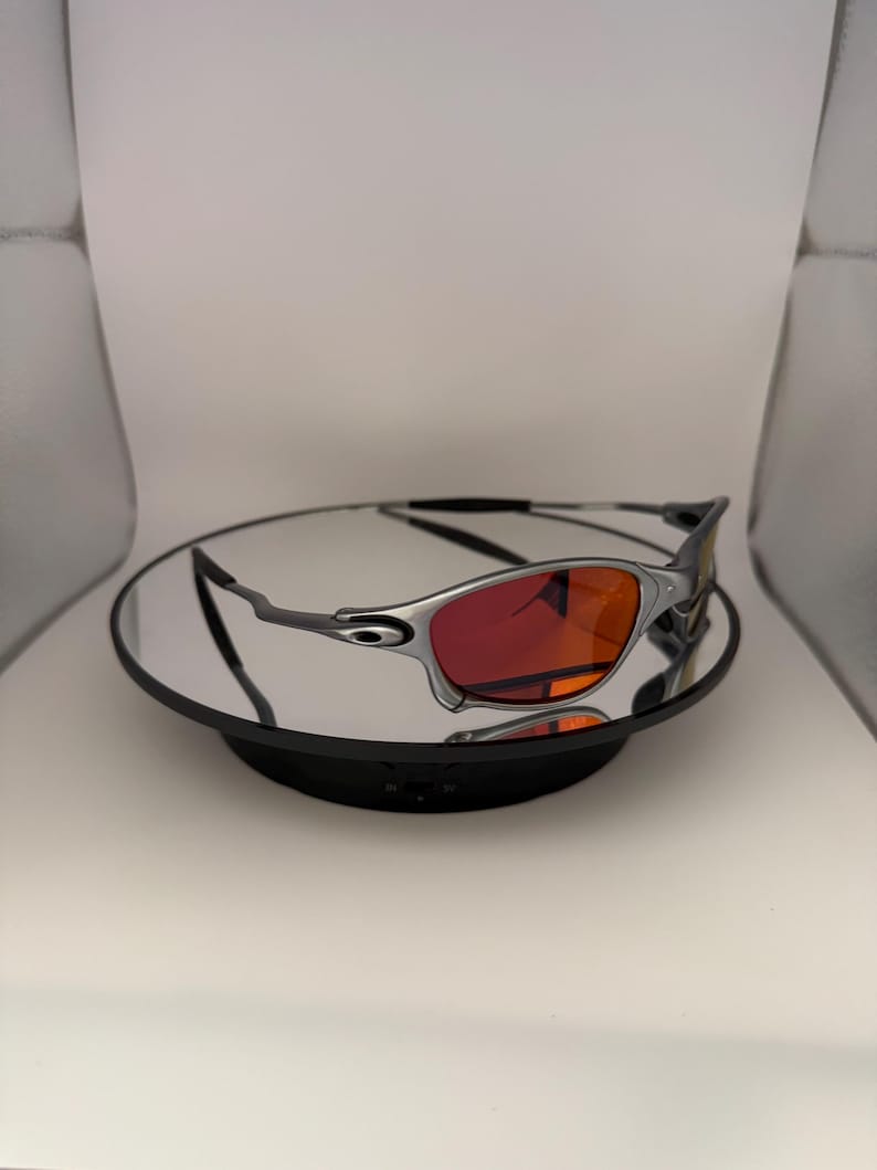 May include: Silver-framed sunglasses with vibrant red lenses. The sunglasses are displayed on a reflective surface, showcasing their sleek design. The lenses offer a striking contrast against the silver frame, creating a modern aesthetic.