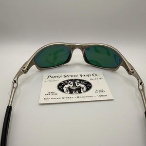 May include: Silver-framed sunglasses with green lenses. The glasses have a sleek, modern design. A business card with the text "Paper Street Soap Co." is visible. The card also includes the text "All Natural" and a phone number.