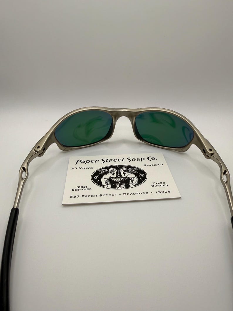 May include: Silver-framed sunglasses with green lenses. The glasses have a sleek, modern design. A business card with the text "Paper Street Soap Co." is visible. The card also includes the text "All Natural" and a phone number.