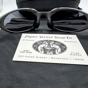 May include: A black and gray pair of sunglasses with a black case. The sunglasses have a unique oval shape and are sitting on a black fabric surface. A business card for Paper Street Soap Co. is also visible in the image. The card includes the company's name, address, phone number, and the text "All Natural" and "Handmade".