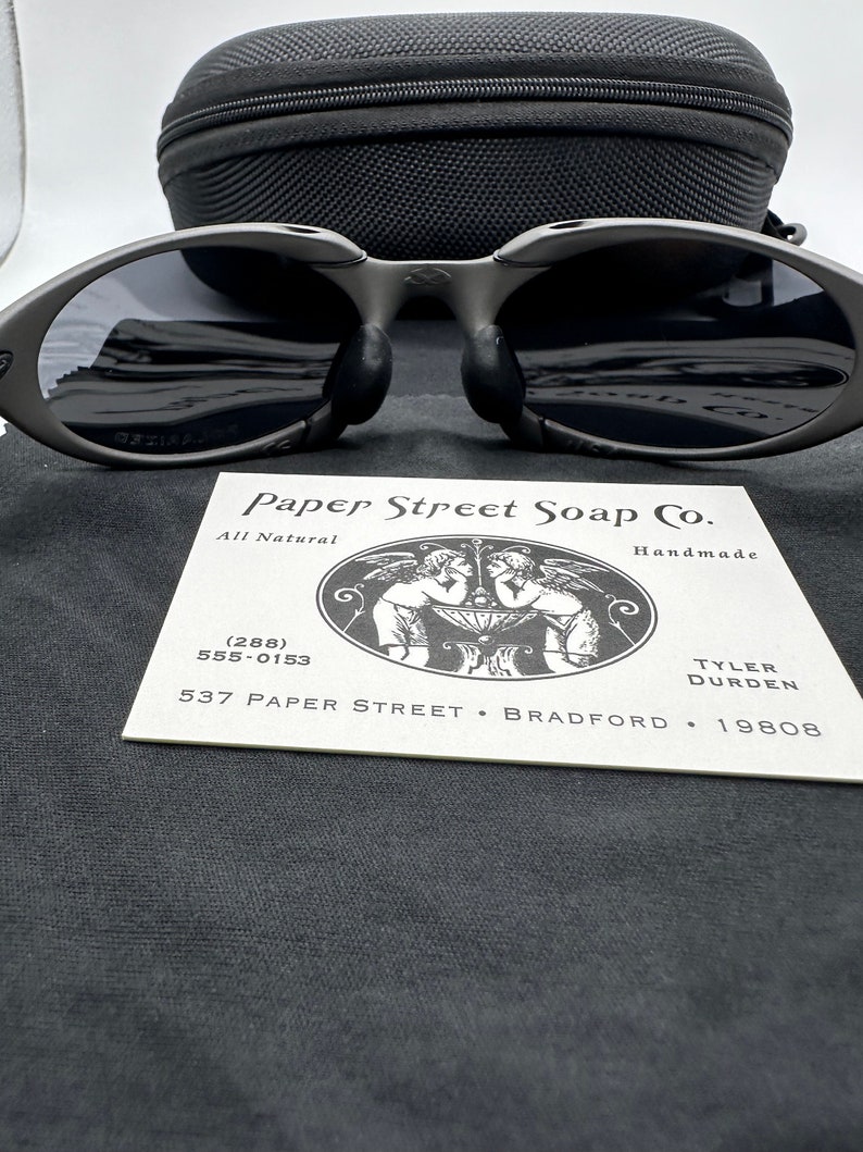 May include: A black and gray pair of sunglasses with a black case. The sunglasses have a unique oval shape and are sitting on a black fabric surface. A business card for Paper Street Soap Co. is also visible in the image. The card includes the company's name, address, phone number, and the text "All Natural" and "Handmade".