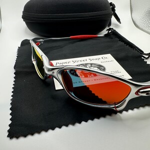 Może przedstawiać: Srebrne okulary przeciwsłoneczne Oakley z pomarańczowymi szkłami i czerwoną opaską. Okulary leżą na czarnym mikrofibrowym ściereczce do czyszczenia z białą kartką, na kt&oacute;rej widnieje napis "Paper Street Soap Co. Handmade" i "Tyler Founder".