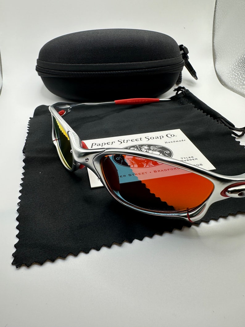 Może przedstawiać: Srebrne okulary przeciwsłoneczne Oakley z pomarańczowymi szkłami i czerwoną opaską. Okulary leżą na czarnym mikrofibrowym ściereczce do czyszczenia z białą kartką, na kt&oacute;rej widnieje napis "Paper Street Soap Co. Handmade" i "Tyler Founder".