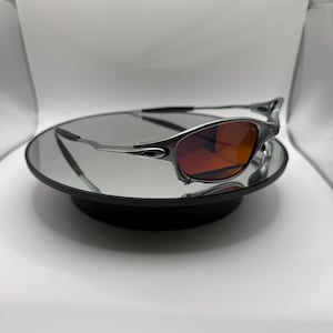 May include: Silver-framed sunglasses with red-tinted lenses. The sunglasses are displayed on a round, black mirrored surface. The lenses have a gradient effect, transitioning from a darker red at the top to a lighter shade at the bottom.