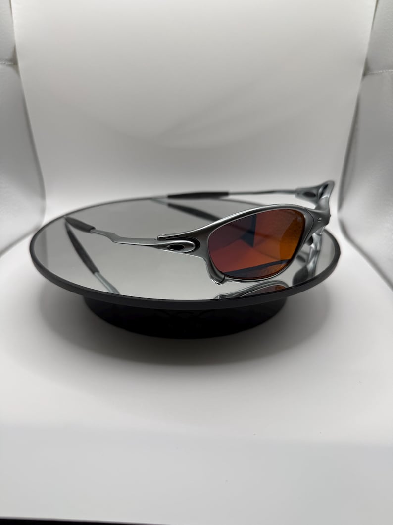 May include: Silver-framed sunglasses with red-tinted lenses. The sunglasses are displayed on a round, black mirrored surface. The lenses have a gradient effect, transitioning from a darker red at the top to a lighter shade at the bottom.