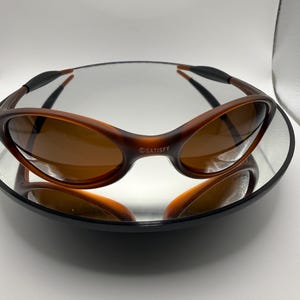 May include: Brown sunglasses with a curved frame and tinted lenses. The sunglasses are resting on a reflective surface. The text "SATISFY" is printed on the frame.