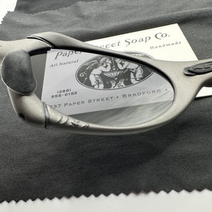 May include: A close-up of a pair of gray sunglasses with a black case. The sunglasses have a unique design and are made by Paper Street Soap Co. The case has the company's logo and contact information.