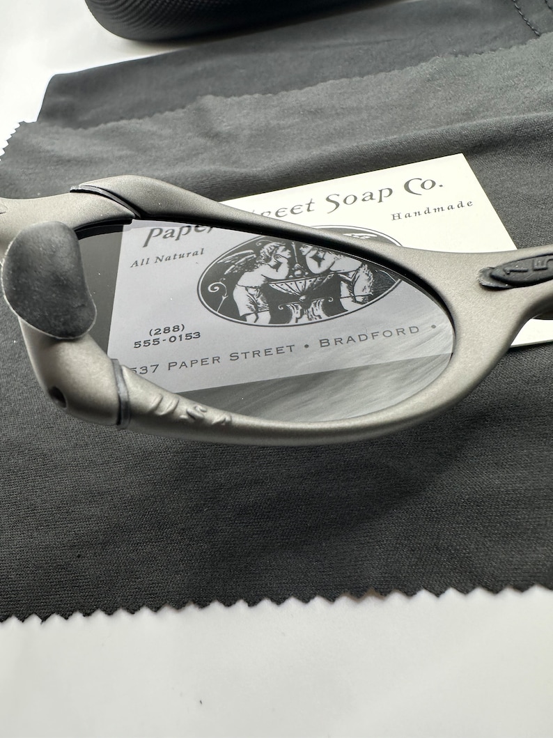 May include: A close-up of a pair of gray sunglasses with a black case. The sunglasses have a unique design and are made by Paper Street Soap Co. The case has the company's logo and contact information.