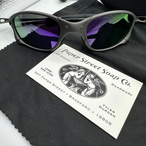 May include: A pair of black sunglasses with green and purple lenses. The sunglasses are resting on a black fabric pouch with a white business card that reads "Paper Street Soap Co. All Natural Handmade Tyler Durden 537 Paper Street Bradford 19808 (288) 555-0153".