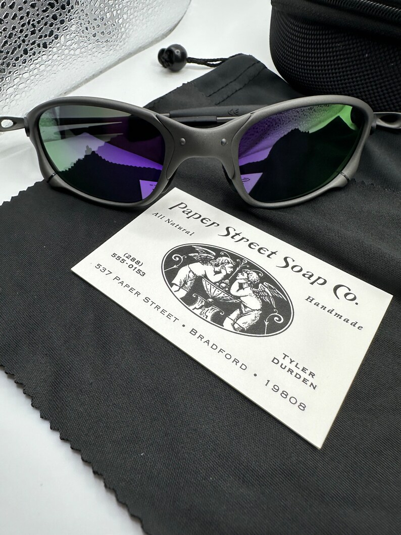 May include: A pair of black sunglasses with green and purple lenses. The sunglasses are resting on a black fabric pouch with a white business card that reads "Paper Street Soap Co. All Natural Handmade Tyler Durden 537 Paper Street Bradford 19808 (288) 555-0153".