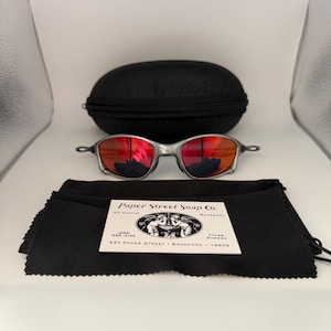 May include: Silver-framed sunglasses with red lenses, a black carrying case, and a black microfiber cleaning cloth. The cloth has a white label with the text "Paper Street Soap Co."
