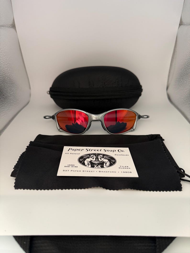 May include: Silver-framed sunglasses with red lenses, a black carrying case, and a black microfiber cleaning cloth. The cloth has a white label with the text "Paper Street Soap Co."