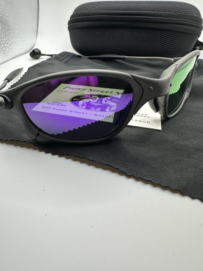 May include: Black sunglasses with green mirrored lenses and a black case. The sunglasses have a purple reflection in the lenses, showing a business card with the text "Paper Street" and "537 Paper Street, Bradford, PA 19808".