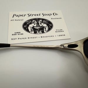 Peut inclure: Lunettes de soleil argent&eacute;es avec des branches et des verres noirs. Une carte de visite de Paper Street Soap Co. est pr&eacute;sente, avec une illustration de ch&eacute;rubins et les mentions "All Natural" et "Handmade".